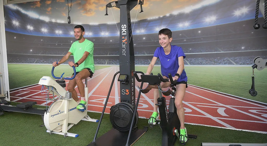 Δύο αθλητές προπονούνται πάνω σε στατικό ποδήλατο. Ο εξοπλισμός προσφέρει καρδιαγγειακή προπόνηση και ενδυνάμωση σε κλειστό χώρο γυμναστηρίου.