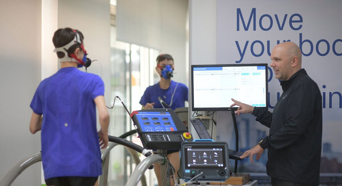 Επαγγελματίας εργοφυσιολόγος παρακολουθεί αθλητή κατά τη διάρκεια εξέτασης VO₂ max πάνω σε διάδρομο τρεξίματος.